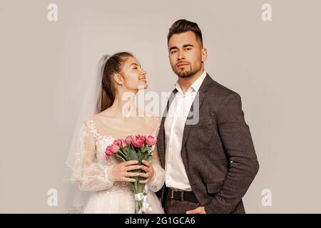 Glückliche Braut, die auf selbstbewussten Bräutigam steht, mit der Hand in der Tasche isoliert auf grau Stockfoto