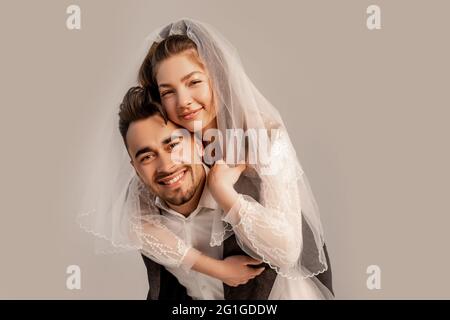 Lächelnde Braut Huckepack auf glücklichen Bräutigam isoliert auf grau mit lila Schatten Stockfoto