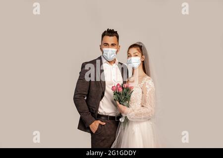 Mann in medizinischer Maske, der mit der Hand in der Tasche in der Nähe der Braut mit Tulpen auf grau isoliert steht Stockfoto