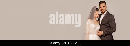 Glücklicher Bräutigam lächelt an der Kamera nahe elegante Braut isoliert auf grau mit lila Schatten, Banner Stockfoto