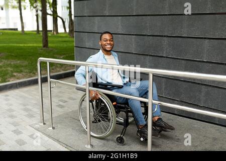Glücklich beeinträchtigter schwarzer Mann im Rollstuhl, der das Gebäude auf einer Rampe im Freien betrat, in voller Länge. Behindertengerechte Stadt Stockfoto