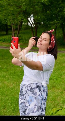 Brünette kaukasische Frau mit Jade Gouache Scraper draußen, Mädchen tun Gouache Gesichtsmassage vor dem Telefon. Vertikales Foto Stockfoto