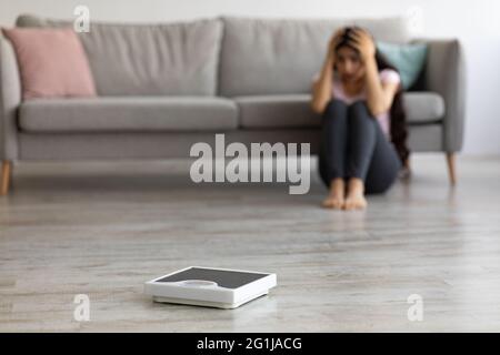 Gestresste junge Inderin, die auf dem Boden sitzt, sich vor Angst den Kopf ergreifend, leere Schuppen ansieht und sich übergewichtig fühlt Stockfoto