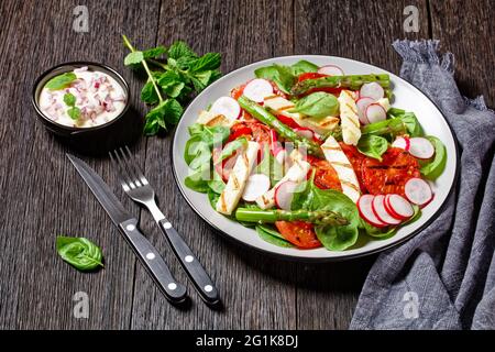 Gegrillte Tomaten, Halloumi-Käse, Spargel, Rettich- und Spinatsalat auf einem Teller auf einem Holztisch mit Mayo-Dressing in einer Schüssel, horizontale Ansicht von Stockfoto