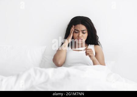 Hohes Fieber, Unwohlsein. Kranke schwarze Frau, die das Thermometer hält und anschaut und die Körpertemperatur im Bett überprüft Stockfoto