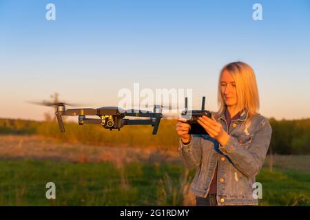 Junge blonde Frau kontrolliert während des Sonnenuntergangs eine Drohne in der Natur. Im Vordergrund ist eine Nahaufnahme-Drohne Stockfoto