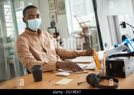 Afrikanischer Geschäftsmann in einer Maske, die an seinem Schreibtisch sitzt Stockfoto