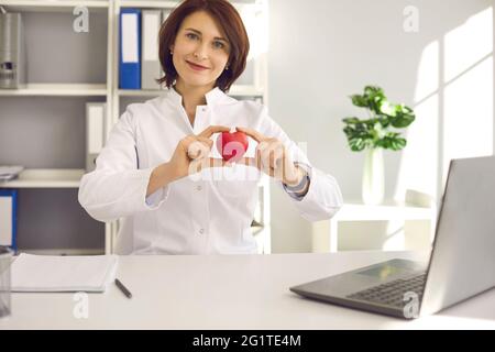 Porträt einer Kardiologin, die ein kleines rotes Herz in der Hand hält, während sie im Büro am Schreibtisch sitzt. Stockfoto