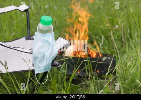 Medizinische Maske, die an einer Wasserflasche hängt. Die Flasche ist im Campingstuhl installiert. In der Nähe brennt ein Feuer in einem Metallgrill. Sommerurlaub während Stockfoto