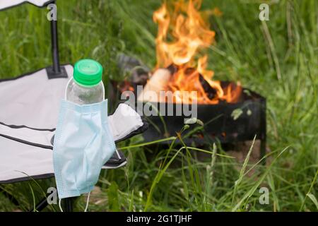 Medizinische Maske, die an einer Wasserflasche hängt. Die Flasche ist im Campingstuhl installiert. In der Nähe brennt ein Feuer in einem Metallgrill. Sommerurlaub während Stockfoto