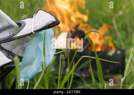 Medizinische Maske, die an einer Wasserflasche hängt. Die Flasche ist im Campingstuhl installiert. In der Nähe brennt ein Feuer in einem Metallgrill. Sommerurlaub während Stockfoto