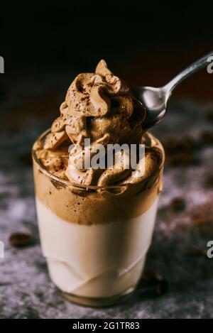 Jemand nimmt einen Löffel Dalgona-Kaffee aus einem Glas, auf einer dunkelgrauen Steinoberfläche vor schwarzem Hintergrund Stockfoto