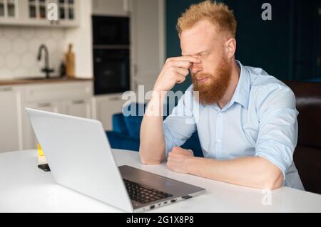 Erschöpfter Rotschopf-Geschäftsmann in elegantem Casual-Shirt, der eine Nasenbrücke hält, fühlt sich die Augen belastet, müde von der Online-Arbeit mit einem Laptop, sitzt müde Kerl mit geschlossenen Augen auf dem Schreibtisch Stockfoto