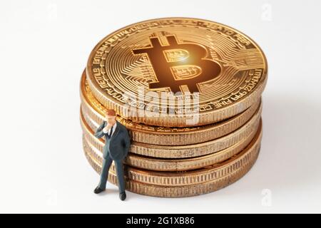 Winzig denkende Geschäftsmann Figur, während sie sich zu glänzendem Bitcoin Stapel auf weißem Hintergrund lehnt. Entwicklung einer Anlagestrategie für Kryptowährungen. Stockfoto