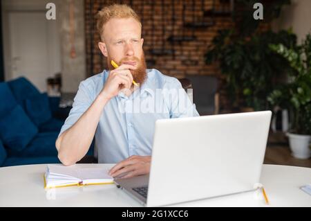Nachdenklicher, moderner rothaariger Geschäftsmann, der nach neuen Ideen sucht, mit einem Laptop im Heimbüro am Schreibtisch sitzt, in Gedanken verloren schaut, Aufgaben löst, plant Stockfoto