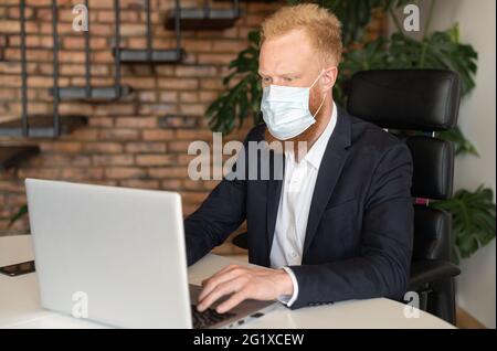 Seriöser rothaariger Geschäftsmann in schickem Freizeitanzug, der eine schützende Gesichtsmaske trägt und einen Laptop-Computer verwendet, der im modernen Büro sitzt, ergreift ein männlicher Mitarbeiter Schutzmaßnahmen gegen Viruserkrankungen Stockfoto