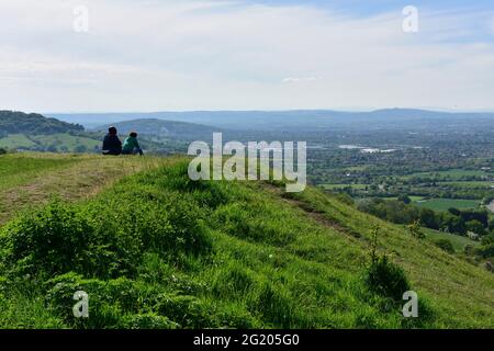 Zwei Personen sitzen auf einer grasbewachsenen Böschung über Severn Val und blicken über Gloucester in Richtung Malvern Hills, Gloucestershire, Großbritannien Stockfoto