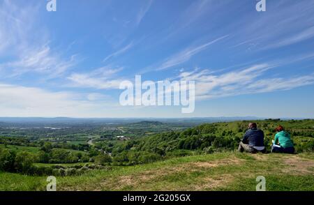 Zwei Personen sitzen auf einer grasbewachsenen Böschung über Severn Val und blicken über Gloucester in Richtung Malvern Hills, Gloucestershire, Großbritannien Stockfoto