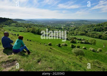 Zwei Personen sitzen auf einer grasbewachsenen Böschung über Severn Val und blicken über Gloucester in Richtung Malvern Hills, Gloucestershire, Großbritannien Stockfoto