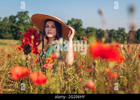 Junge Frau pflücken Strauß von Mohnblumen Blumen Wandern im Sommerfeld genießen Blick auf die schöne Landschaft trägt Vintage Strohhut Stockfoto