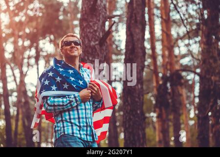 Glücklicher Mann, der bei Sonnenuntergang die USA-Flagge im Park hält. Feiern des Unabhängigkeitstages von Amerika. Juli. Mann, der Spaß hat Stockfoto