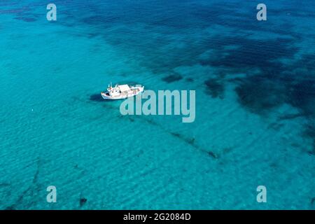 Fischerboot auf türkisblauem Meereshintergrund. Luftdrohnenansicht. Blau und weiß traditionelle Trawler, die im ägäischen gewellten Wasser festgemacht sind, sonniger Tag. Stockfoto