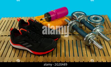 Gym-Set aus schwarzen Sportschuhen, Hanteln zum Trainieren, gelbem Handtuch und roter Flasche mit Wasser auf Holzboden mit blauem Hintergrund Stockfoto