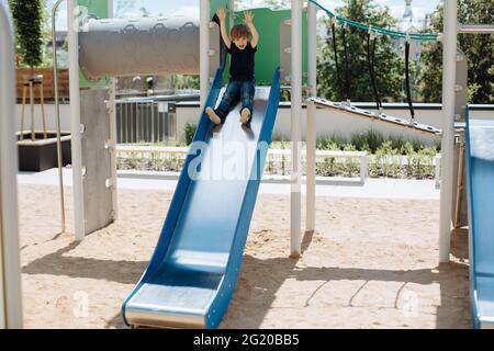 Junge 3 Jahre alt spielt auf Spielplatz im Hof im Freien. Glücklich fährt er mit hochgekommenen Händen bergab. Stockfoto