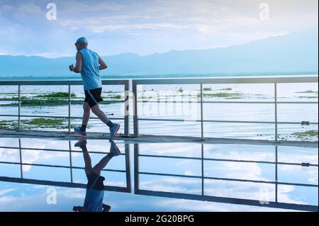 Bewegungsunschärfe. Ein asiatischer älterer Mann in Fitnesskleidung läuft in einem Park entlang eines Sees bei Sonnenaufgang, blaue Berge verschwommen im Hintergrund. Stockfoto