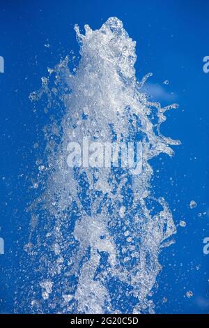 Ein Wasserstrahl, der unter Druck eines Brunnens gegen einen blauen Himmel gesprüht wird, der von Sonnenlicht aus der Nähe beleuchtet wird. Stockfoto
