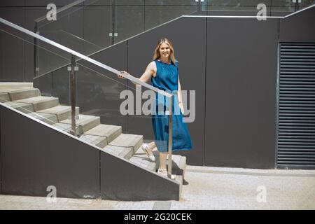 Junge Frau in legerer Kleidung geht die Treppe hinunter, schaut in die Kamera, kopiert den Raum. Stockfoto