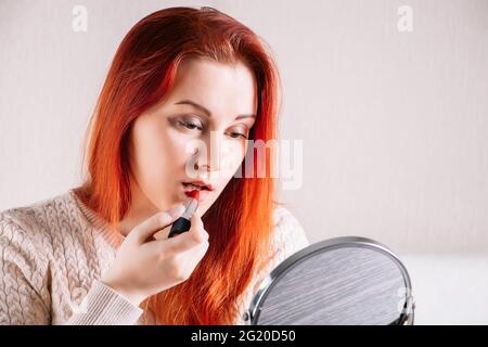 Junge rothaarige Frau schminkt sich und sieht in den Spiegel. Weibchen trägt Lippenstift. Stockfoto