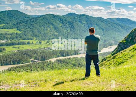 Ein Mann steht auf einem Berg und blickt nachdenklich auf ein Bergtal mit einem Fluss Stockfoto