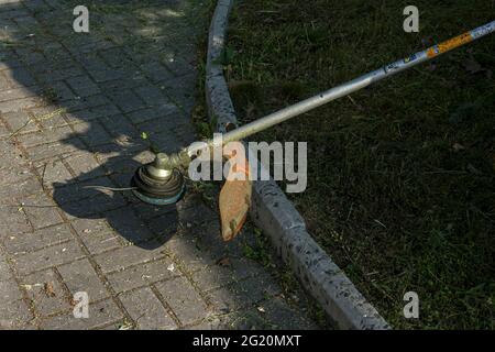 Ein Arbeiter im Overall mit einem Trimer in den Händen mäht das Gras auf dem Rasen und die Wege im Park. Gebietsbetreuung. Stockfoto