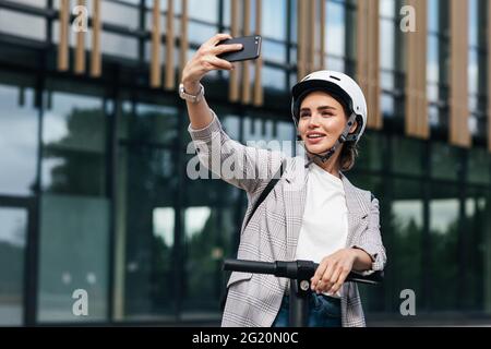 Junge schöne Frau in einem weißen Fahrradhelm, die Selfie nimmt, während sie auf einem elektrischen Motorroller in der Stadt steht Stockfoto