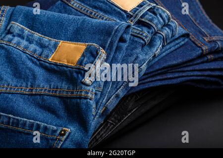 Jeans-jeans-Hose im Stapel gefaltet mit leerem braunem Tag-Mockup-Label. Lässige blaue Jeans im Stapel auf schwarzem Hintergrund. Stockfoto