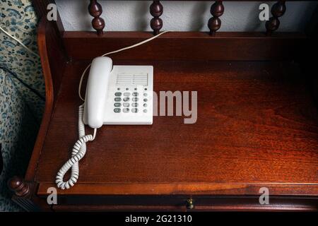 Altes weißes Telefon auf einem Vintage-Nachttisch aus braunem Holz in der Nähe des Bettes im Schlafzimmer mit antikem Design und Platz für Kopien Stockfoto