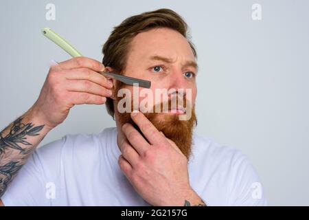 Ein ernsthafter Mann mit Klinge konzentriert sich darauf, seinen Bart zu schneiden Stockfoto