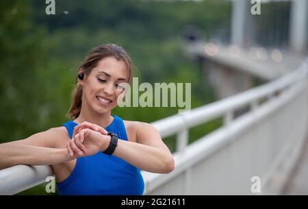 Weibliche Athletin, die eine intelligente Uhr zum Laufen aufsetzt. Junge athletische Frau, die sich beim Training auf dem Brückengeländer stützt. Stockfoto