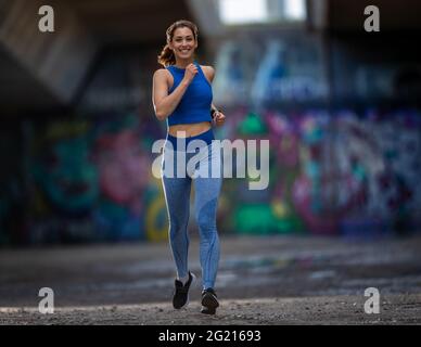 Fit junge Frau Joggen im städtischen Bereich. Athlet Speed Walking lächelt mit einer mit Graffiti bedeckten Wand im Hintergrund. Stockfoto