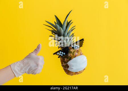 Die Hand im Handschuh zeigt das Zeichen „Daumen hoch“. Schwebender Ananas mit witzigem Gesicht, Brille und schützender medizinischer Maske. Reise-Coronavirus-Konzept Stockfoto