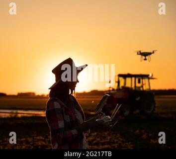 Weibliche Agronomin fliegende Drohne mit Controller-Innovation in der Landwirtschaft. Junger Bauer steht auf dem Feld mit Traktor im Hintergrund bei Sonnenuntergang. Stockfoto