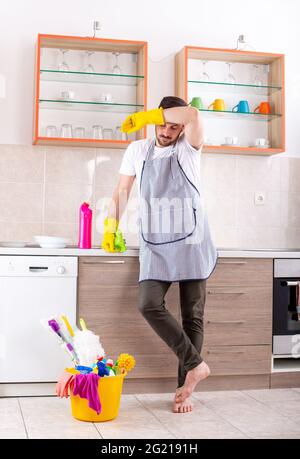 Attraktiver Mann, der in der Küche steht und seine Stirn wischt. Junger Mann, der die Küche mit Wischmopp und Reinigungsmittel putzt und Schutzhandschuhe trägt. Stockfoto