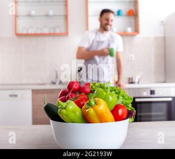 Schüssel mit Paprika Gurken Tomaten Salat Radieschen im Fokus. Lächelnder junger Mann trinkt Kaffee verschwommen und lehnt sich an den Tresen. Stockfoto
