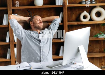 Friedliche Ruhe zufriedener junger kaukasischer Manager oder Freiberufler, der an einem Tisch im Büro sitzt, Pause von der Arbeit macht, seine Hände hinter den Kopf legt und seine Augen schließt, lächelt und chillt Stockfoto