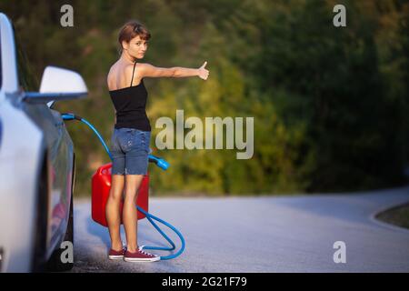 Mädchen, das neben einem Elektroauto steht, hält ein Ladekabel und einen Gassolinenkanister, während es mit dem Anhalter unterwegs ist. Stockfoto