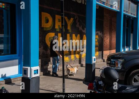 Ein älterer Mann, der blaue Jeans und ein schwarzes T-Shirt trägt, geht mit seinem Hund, einem Corgi-Mix, auf dem Bürgersteig an einem Wandbild in der Innenstadt von Aspen, Colorado, vorbei. Stockfoto