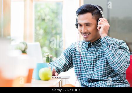 Junger asiatischer Mann, der Kopfhörer trägt, während er im Büro ein Online-Video auf einem Laptop ansieht Stockfoto