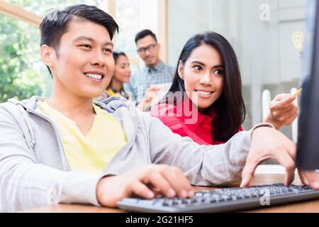 Fröhlicher junger Mitarbeiter, der an einem Desktop-PC arbeitet und dabei hilfreichen Hinweisen seiner erfahrenen Kollegin im Büro folgt Stockfoto