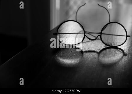 Eine altmodische Vintage-Brille mit Drahtrahmen, durch die Licht auf einem alten Holztisch in Schwarz und Weiß mit geringer Tiefe durchscheint Stockfoto